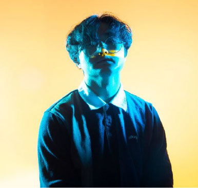 A young adult with medium-length dark hair stands against a bright yellow background. Their face and shirt are illuminated with dramatic blue lighting, creating a high-contrast, artistic portrait. They wear glasses and a collared shirt, looking slightly upward with a calm, neutral expression.
