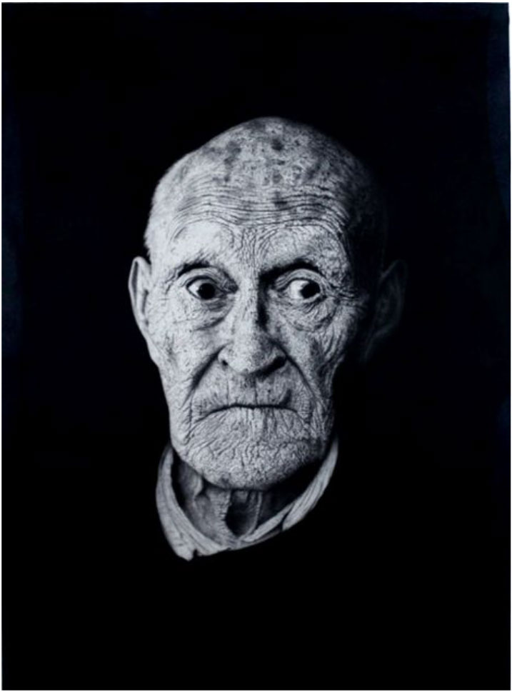 A highly detailed black-and-white portrait of an elderly person with deep wrinkles and expressive, wide-set eyes. Their face is illuminated against a dark background, highlighting the texture of their skin and the folds around their eyes and mouth. The person wears a collared shirt beneath a dark garment, creating a strong contrast with the dramatic lighting.