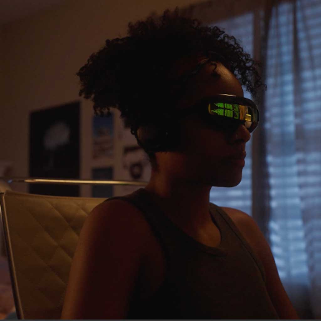 In a dark room, a woman wearing dark sunglasses sits facing right. Reflected in her sunglasses is a screen displaying code. She sits in front of a window with light blue curtains, on a mid-century modern chair.