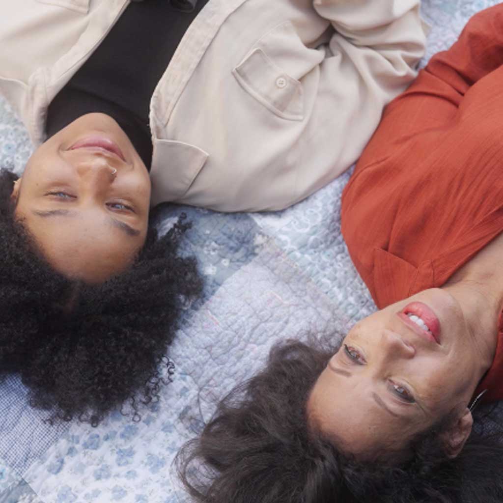 A young woman and an older woman lie on a quilted light blue cover, smiling as they look up at the ceiling. The young woman wears a black T-shirt with a cream-colored over shirt and a nose ring; the older woman wears burnt orange clothes.