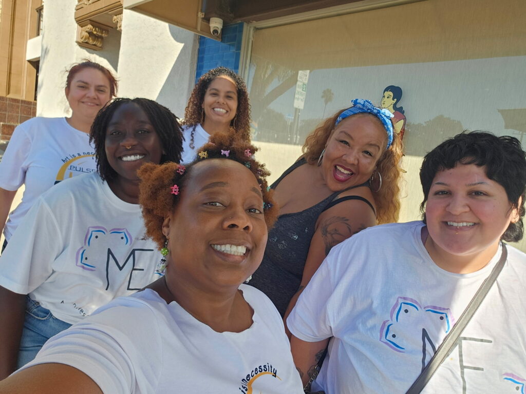 Healing Forward team members smiling together outdoors, wearing white T-shirts, with natural hair styles, headbands, and colorful accessories during a community healing event.